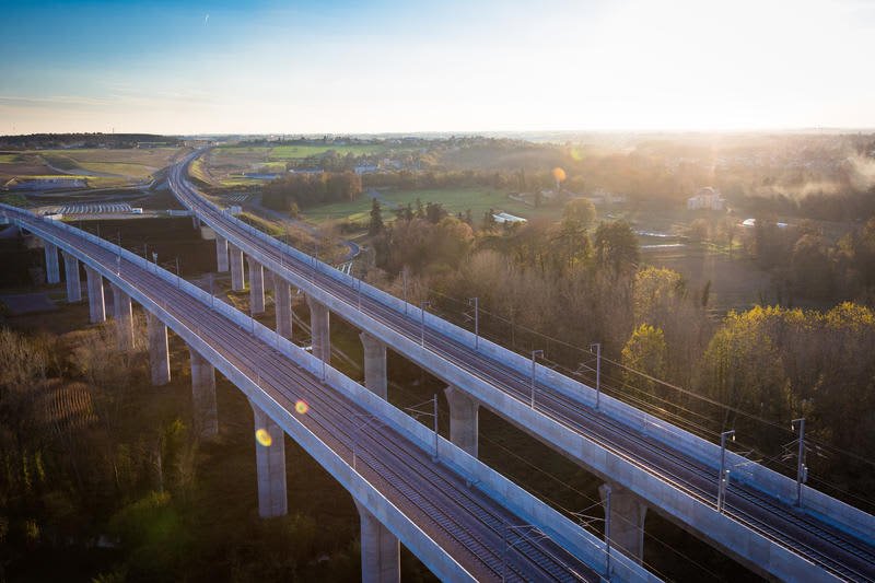 Two parallel elevated railway viaducts on concrete pillars stretch across a rural landscape of trees and fields at sunrise.