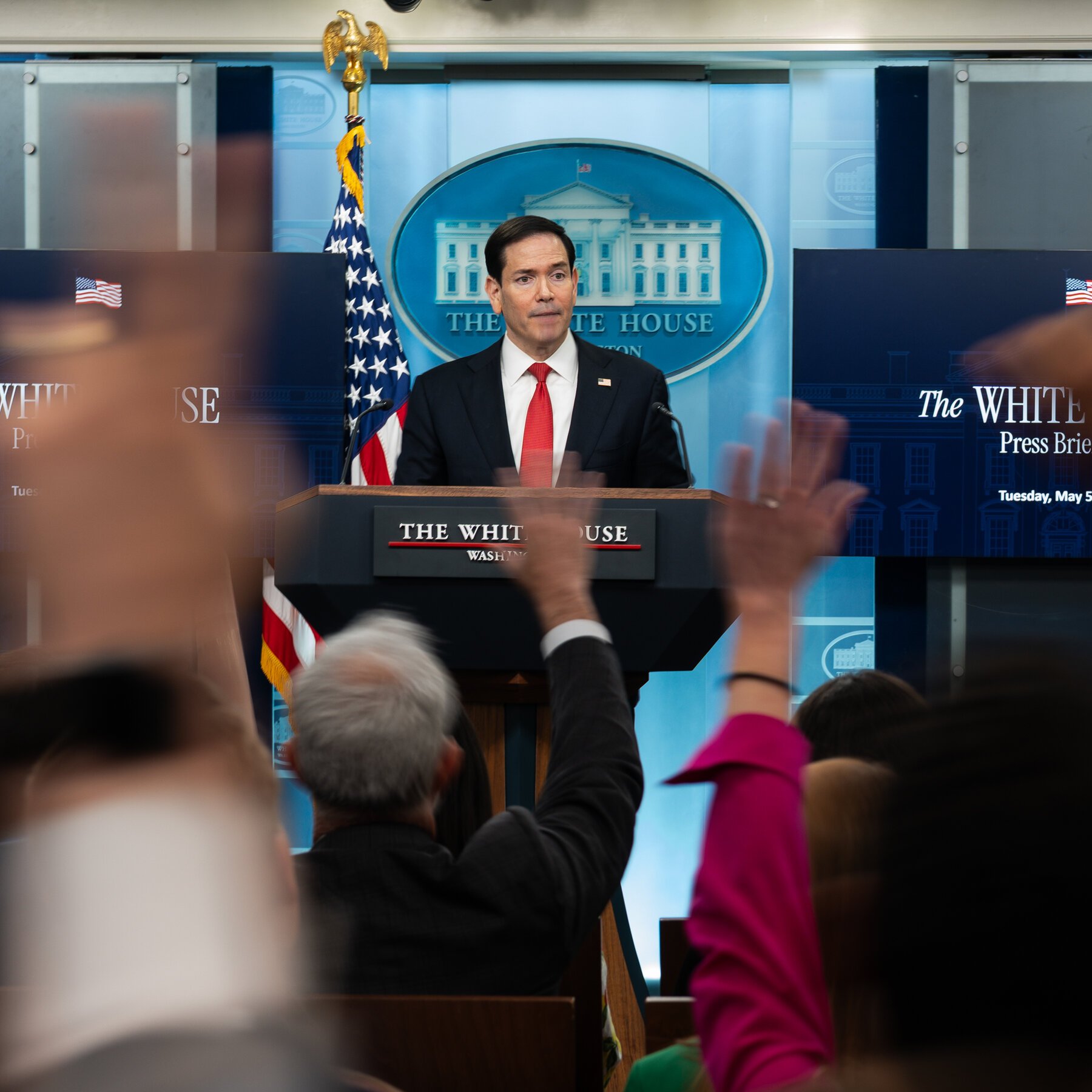 A man in a dark suit and red tie stands at a White House podium as audience members raise their hands, likely to ask questions.