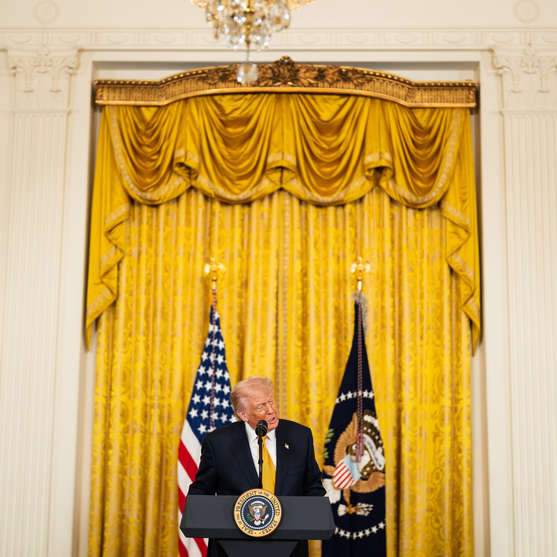 A man in a dark suit and yellow tie speaks at a podium bearing the presidential seal, flanked by the American flag and another flag, against gold drapery.