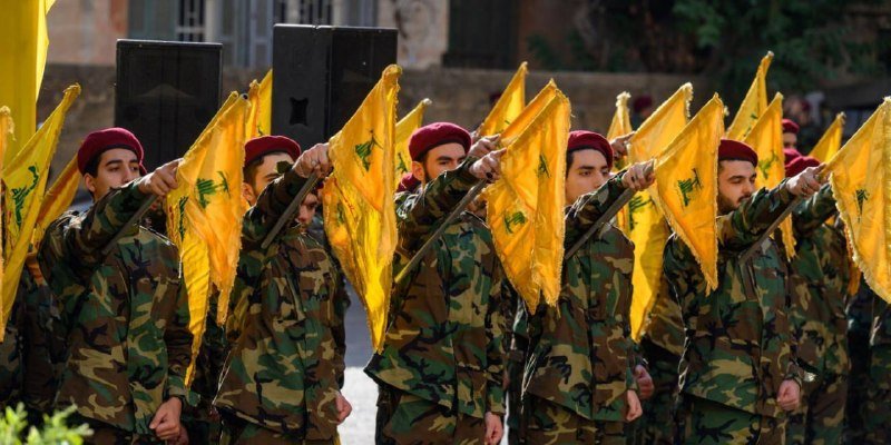 Soldiers in camouflage uniforms and maroon berets march in formation, each holding a yellow flag bearing a green emblem, during an outdoor parade.