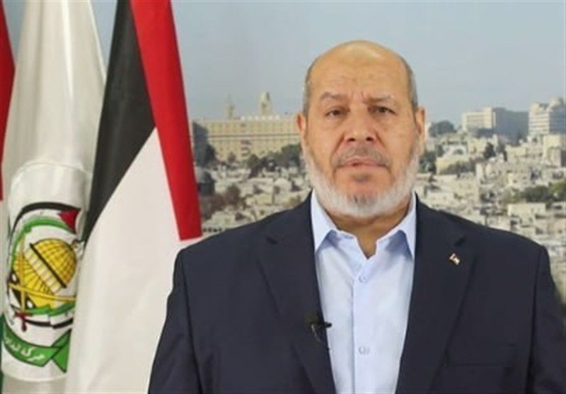A bald, gray-bearded man in a dark suit speaks in front of a flag, with a cityscape visible in the background.