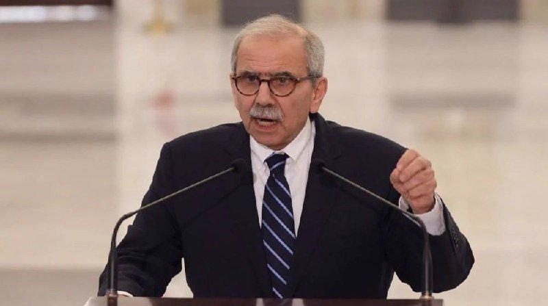 A man in a dark suit, striped tie, and glasses speaks at a podium with microphones, gesturing with one hand.