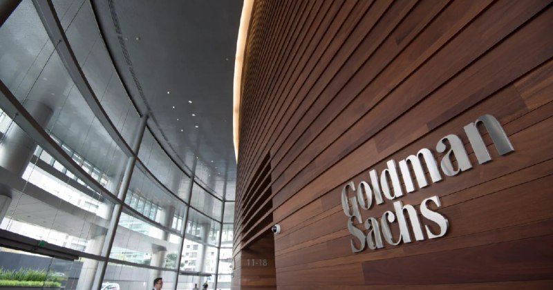 Goldman Sachs signage on a wood-paneled wall inside a modern glass-walled building lobby.