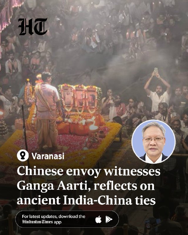A Hindu priest conducts a flower-decorated fire ritual before a seated crowd at night, with smoke rising and an attendee holding up a phone to record.