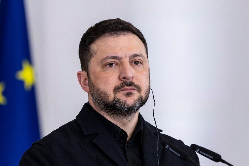 A bearded man in a dark jacket wears an earpiece while seated in front of a European Union flag and Sennheiser microphones.