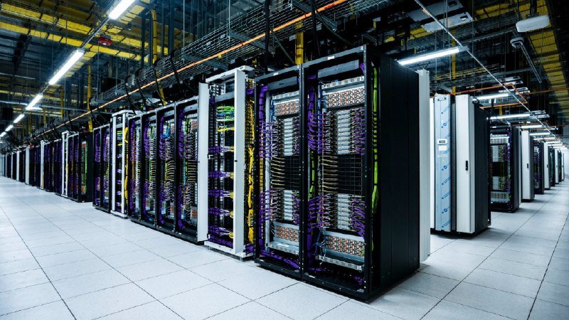 Rows of tall server racks with bundled cables fill a large, brightly lit data center facility.