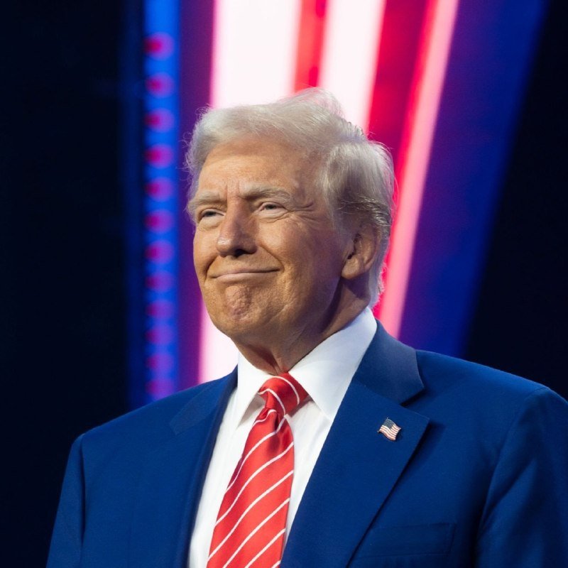 An older man with light hair, wearing a blue suit, red striped tie, and an American flag lapel pin, smiles on a stage lit with red, white, and blue lights.