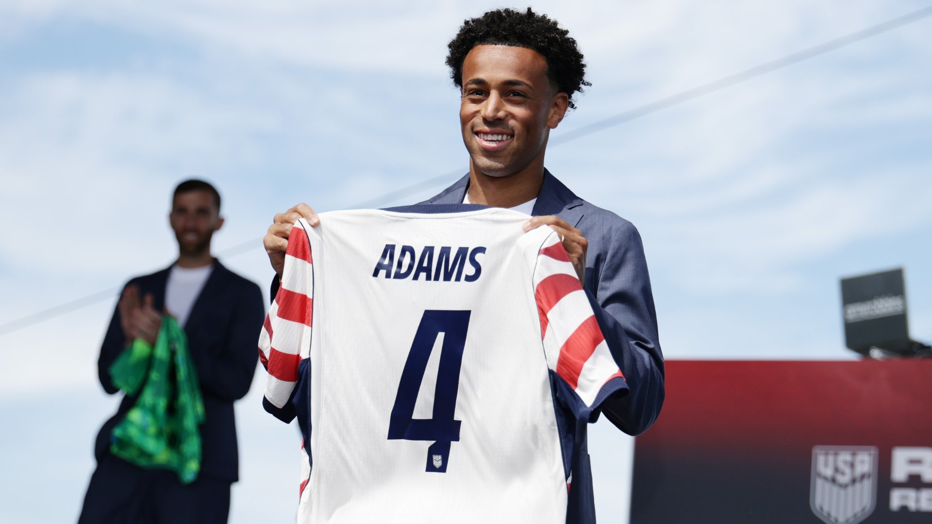 A smiling man in a blue suit holds up a white jersey reading "ADAMS" and "4" with red-and-white striped sleeves, standing before a USA-branded backdrop while another man claps behind him.
