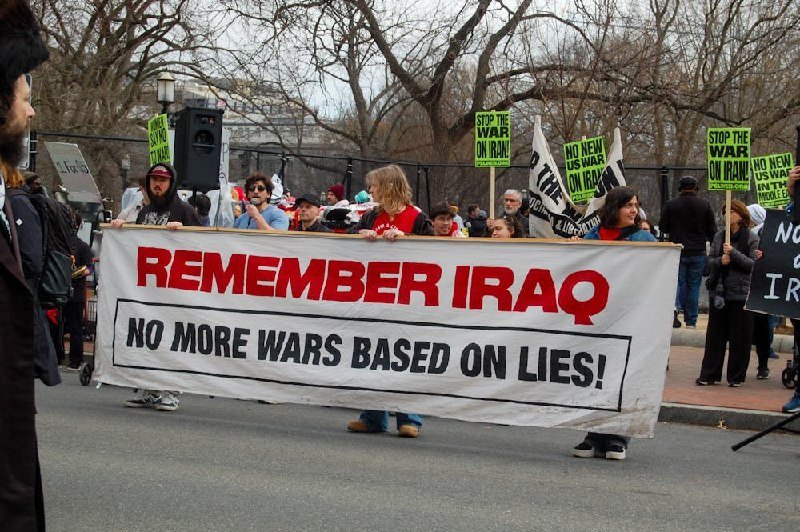 Demonstrators hold a large banner reading "REMEMBER IRAQ - NO MORE WARS BASED ON LIES!" while carrying protest signs during an outdoor rally.