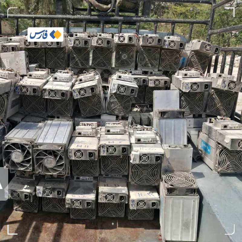 Numerous used cryptocurrency mining machines stacked on a metal flatbed trailer outdoors, with a Persian-language logo visible in the corner.