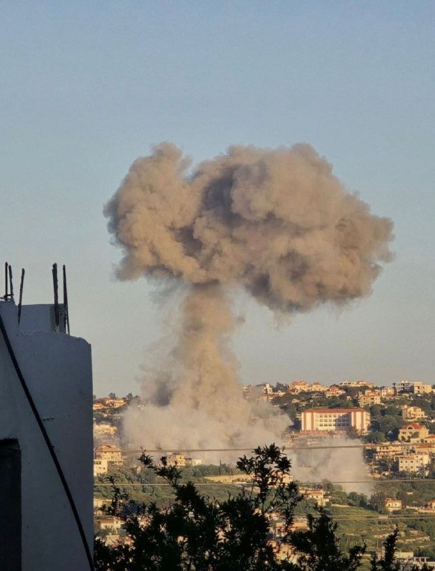 A large mushroom-shaped plume of smoke rises above a hillside town with buildings and trees, viewed past a white structure with exposed rebar in the foreground.