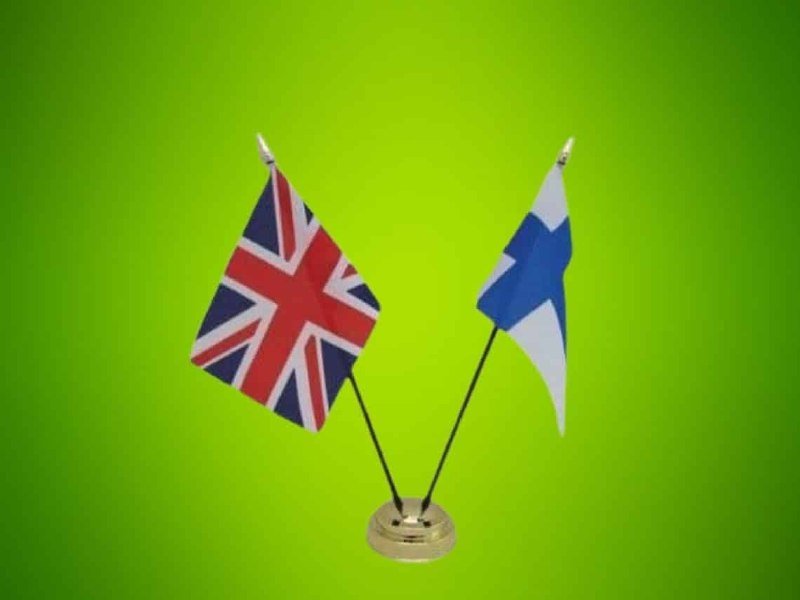 Two small desktop flags—one featuring the Union Jack design and the other a white flag with a blue cross—stand crossed on a golden base against a green background.