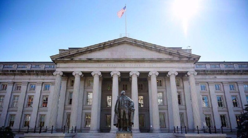 The U.S. Treasury Department building features a neoclassical facade with tall columns, an American flag atop the roof, and a bronze statue standing in front under a sunny sky.