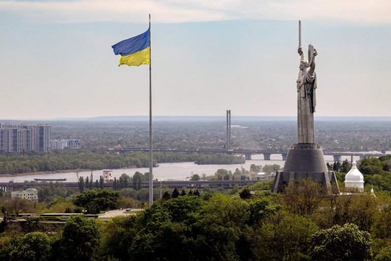 A large blue and yellow flag flies at half-mast beside a tall monument holding a sword, overlooking a river, bridge, and sprawling cityscape.