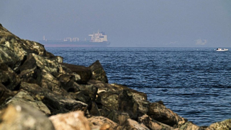 A large cargo ship sails on the horizon beyond a rocky shoreline, while a small motorboat moves across the water to the right.
