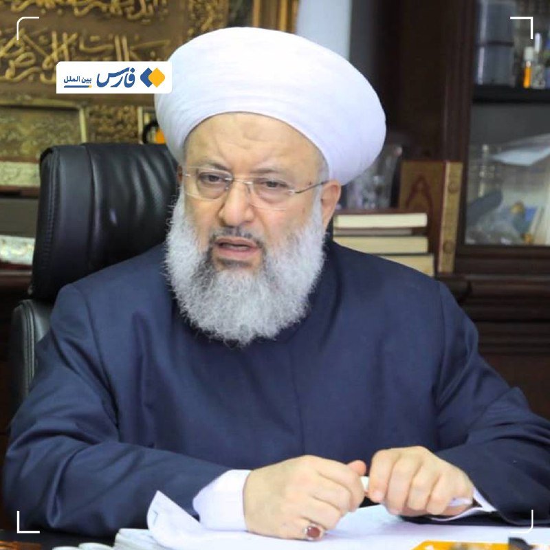 A bearded man in a white turban and dark robe sits at a desk holding papers, with an Arabic-script logo in the upper left.