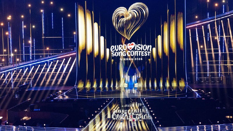 An illuminated stage displays a golden heart sculpture above the "Eurovision Song Contest Vienna 2026" logo, with vertical light beams and a reflective floor.