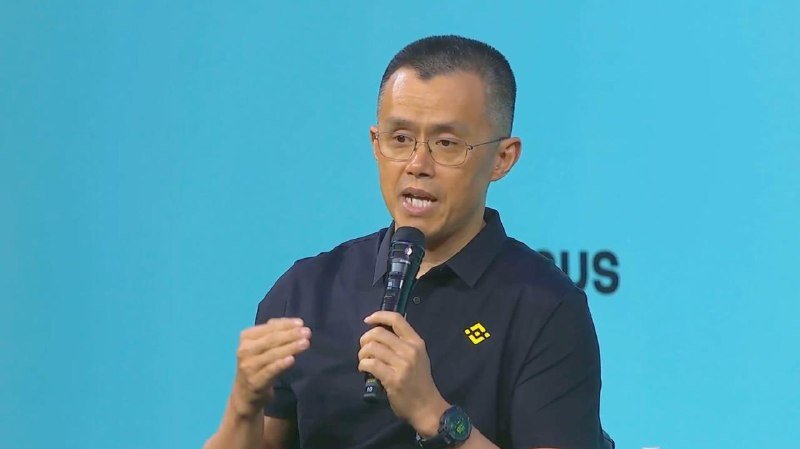 A bespectacled man in a black polo shirt speaks into a microphone while gesturing with his hand against a light blue background with partial text visible.