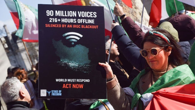 A woman at a protest holds a placard reading "90 MILLION VOICES, 216+ HOURS OFFLINE, SILENCED BY AN IRGC BLACKOUT — WORLD MUST RESPOND, ACT NOW," surrounded by people waving flags.