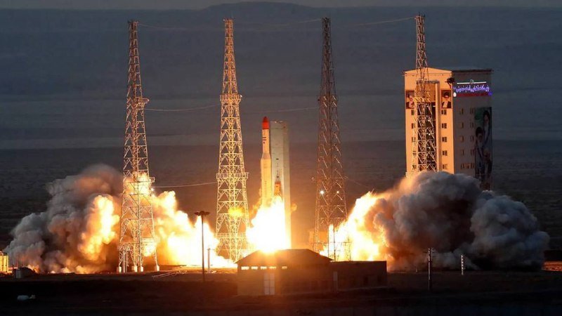 A rocket lifts off from a launch pad flanked by four tall towers, with bright flames and billowing smoke at its base.
