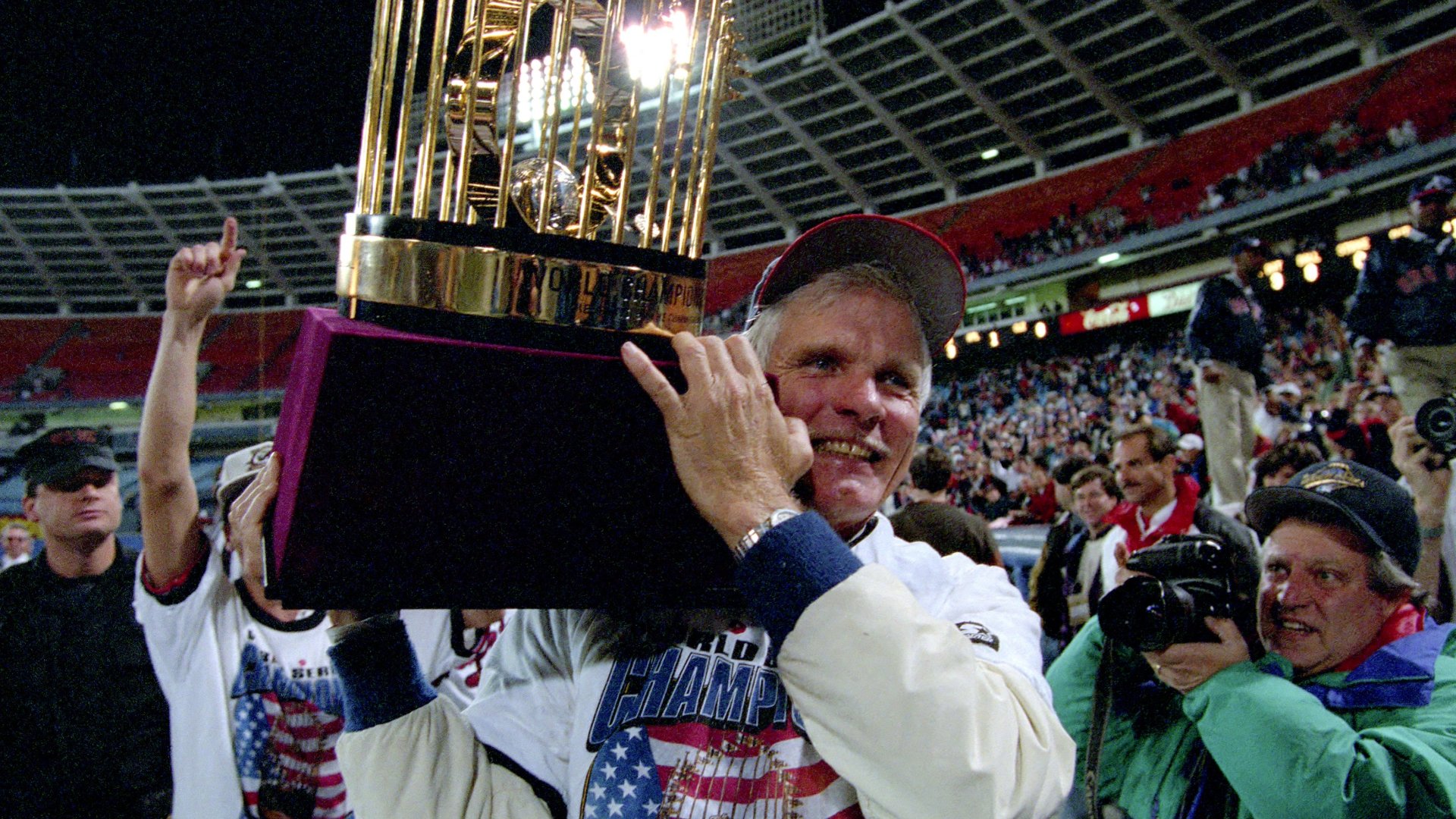 A smiling man in a championship jacket hoists a gold baseball trophy above his head in a crowded stadium, surrounded by photographers and cheering fans.