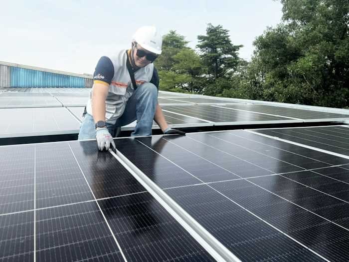 A worker wearing a hard hat, sunglasses, and gloves kneels on a rooftop while installing or inspecting solar panels.
