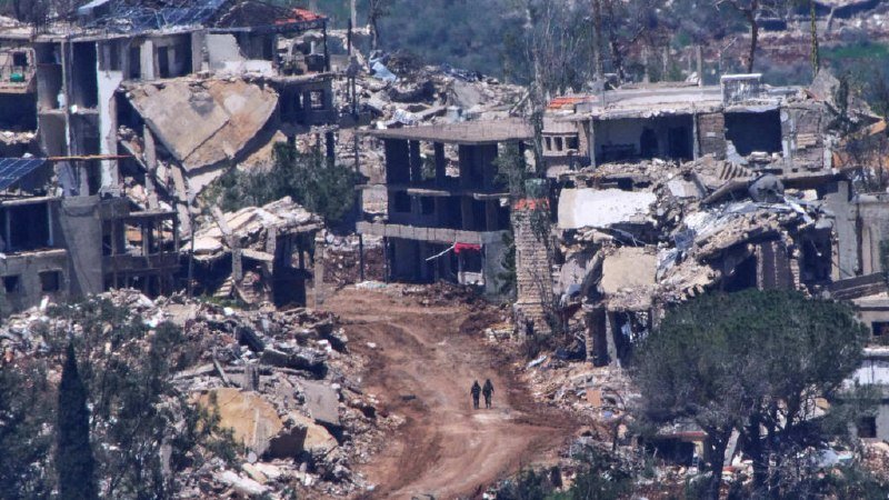 Two people walk down a dirt road through a heavily destroyed urban area with collapsed and damaged buildings surrounded by rubble.