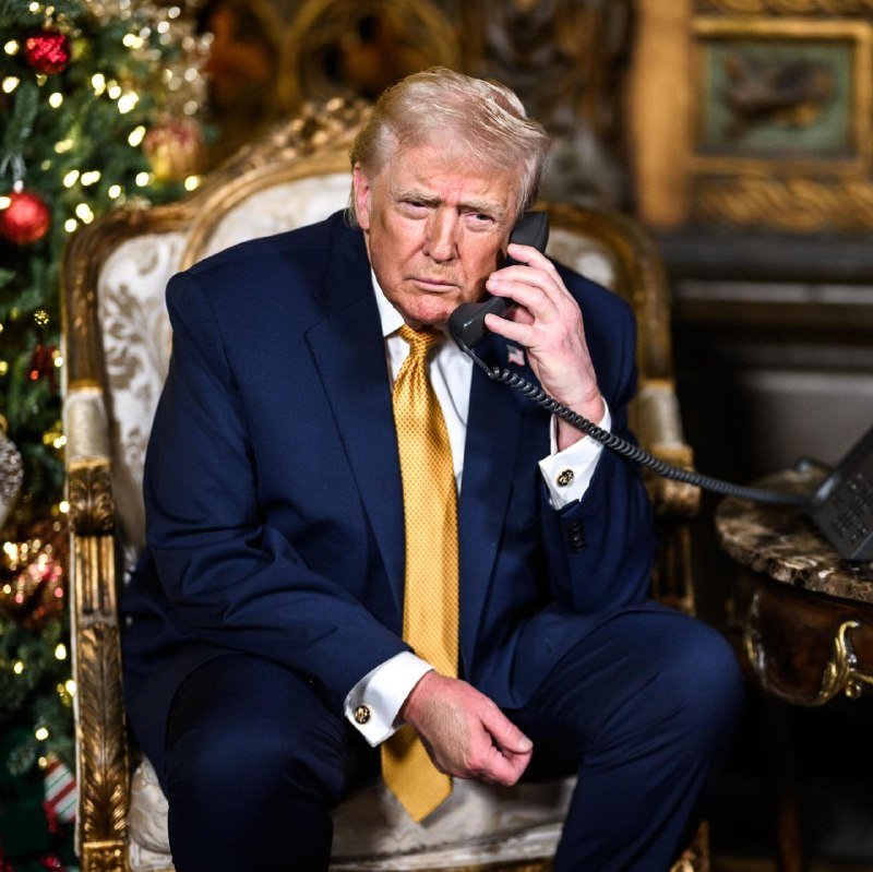 A man in a dark blue suit and gold tie sits in an ornate gold chair, holding a black telephone receiver to his ear next to a decorated Christmas tree.