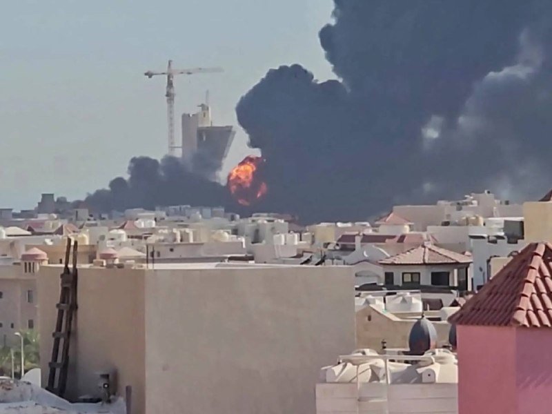 Thick black smoke and bright orange flames rise from behind a row of low-rise buildings in a sunny urban area, with a construction crane visible in the background.