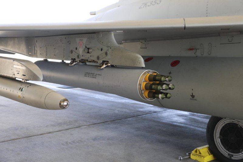 A gray military aircraft marked "ZK353" sits on a tarmac with a rocket pod of green-tipped projectiles mounted under its wing.