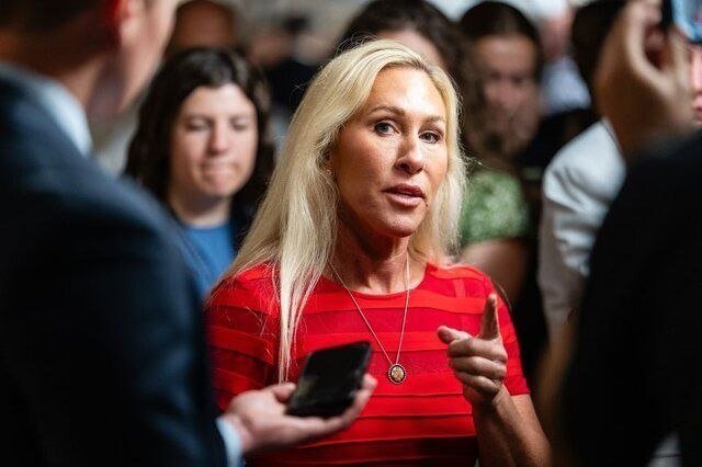 A blonde woman in a red striped top speaks while gesturing, holding a smartphone, surrounded by people in a crowded indoor setting.