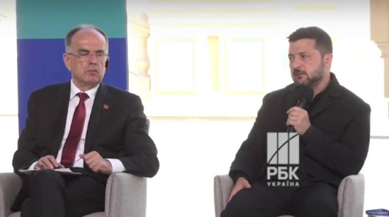 Two men in dark suits sit in chairs during a panel discussion, with one speaking into a microphone; an "РБК Україна" logo appears on screen.