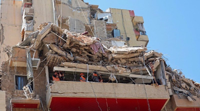 Rescue workers in orange vests stand amid the rubble of a partially collapsed multi-story building, searching through debris and exposed rebar under a clear blue sky.