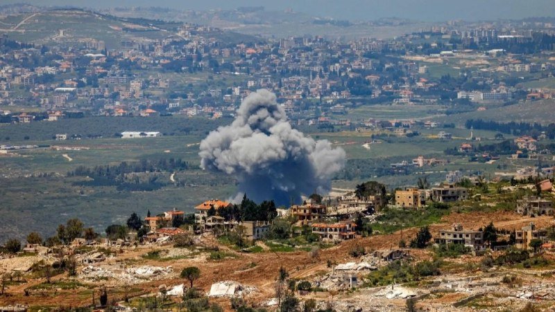 A large plume of gray smoke rises above a hillside village of densely packed buildings, with a broader town and green valleys visible in the background.