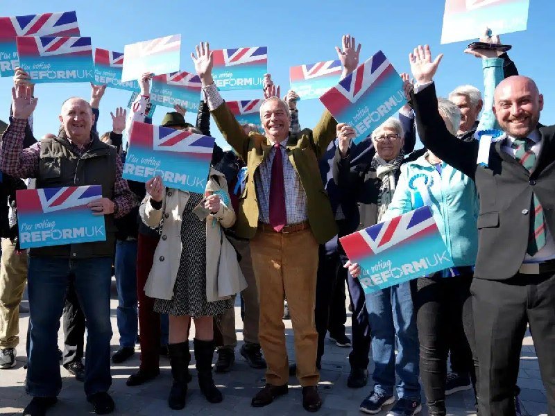 A smiling group of people stand outdoors holding up blue placards reading "I'm voting REFORM UK" alongside Union Jack flags against a clear sky.