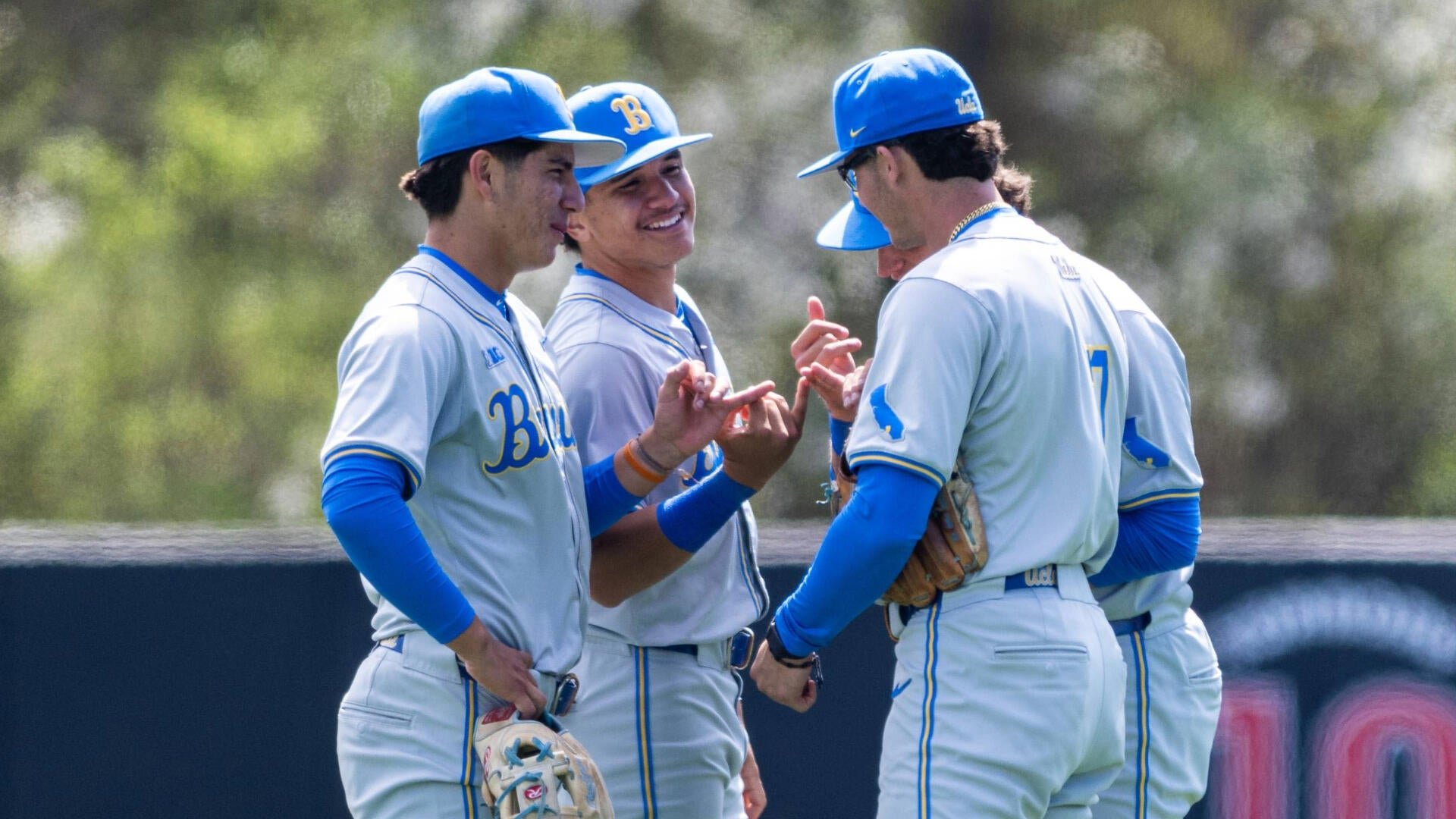 UCLA baseball players in gray and blue uniforms gather on the field, smiling and conversing near the dugout wall.