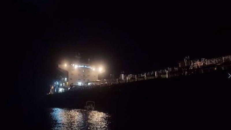 A large illuminated cruise ship is docked at a port at night, with its lights reflecting on the dark water below.