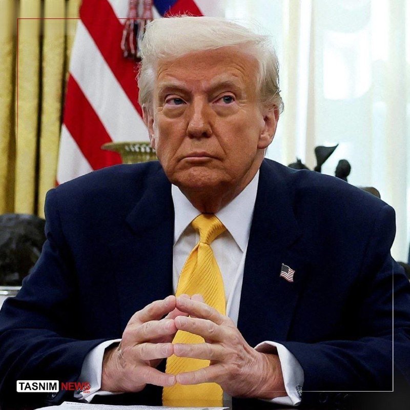 An older man with blonde hair in a dark suit and yellow tie sits with clasped hands in front of an American flag and yellow curtains.