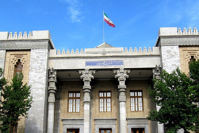 The Iranian flag flies above a stone building featuring ornate columns, arched windows, and a sign with Persian script, flanked by green trees under a clear blue sky.