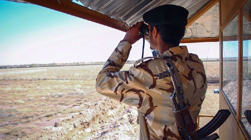 A soldier in desert camouflage uniform and black beret looks through binoculars from a guard tower overlooking an arid landscape, with a rifle slung across his back.