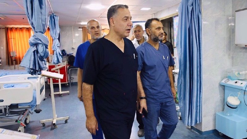 A group of men in medical scrubs walk through a hospital ward with beds and medical equipment visible.