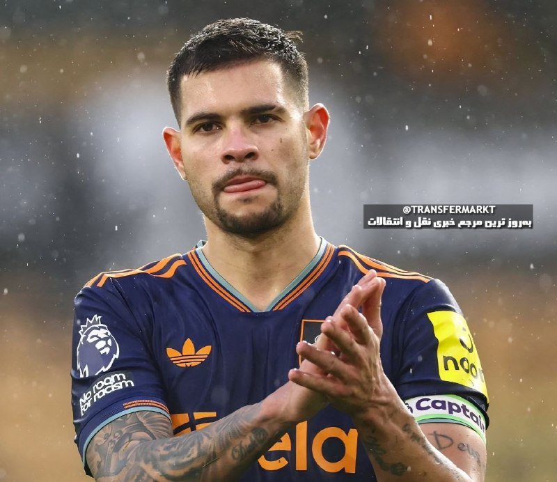 A man in a dark blue soccer jersey with orange trim and a captain's armband claps his hands, with Arabic text visible nearby.