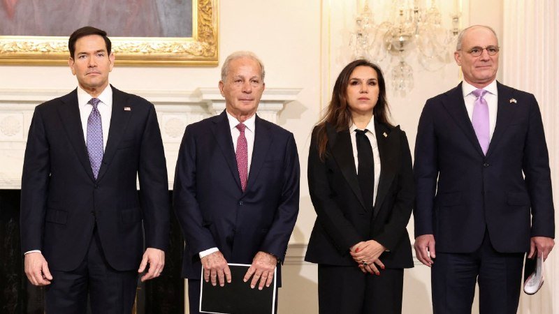 Four people in formal business attire stand side by side in an ornate room featuring a chandelier and framed painting.