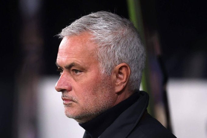 A gray-haired man in a dark coat looks to the side with a serious expression against a blurred dark background.