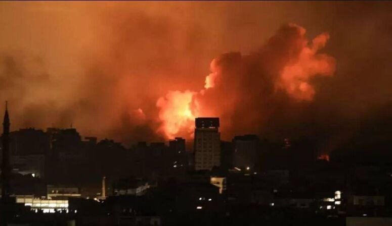 Large flames and thick smoke rise above a silhouetted city skyline at night.