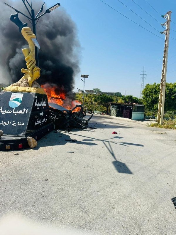 Thick black smoke billows from a burning vehicle beside a monument with Arabic script, on a paved street with power lines and a transmission tower.