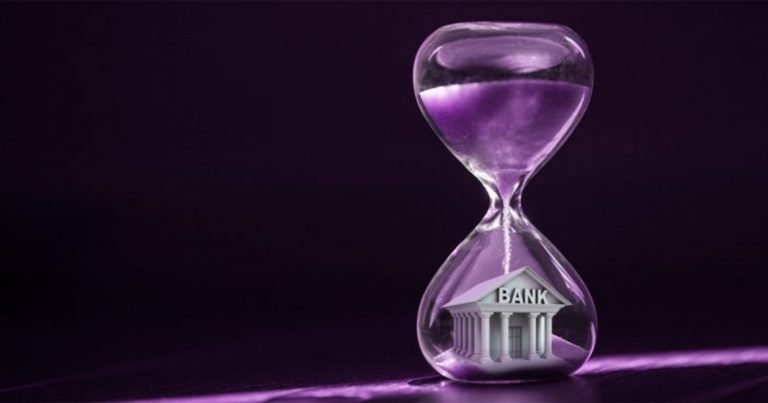 A clear hourglass with purple sand pouring over a small bank building model sits against a dark purple background.