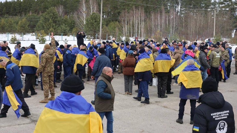 A crowd of people draped in blue and yellow Ukrainian flags gathers outdoors in winter, with some wearing military fatigues and one person holding up a phone to photograph the assembly.