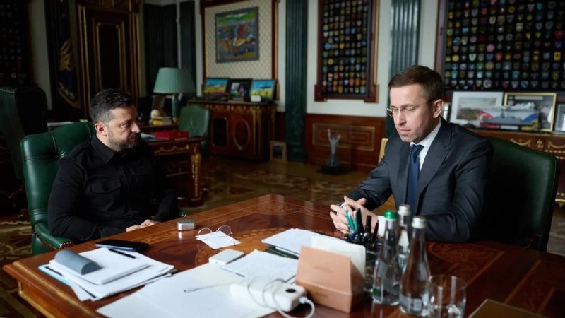 Two men sit across from each other at a wooden desk in an ornate office, one in a dark shirt and the other in a suit gesturing while speaking, with documents and water bottles on the table.