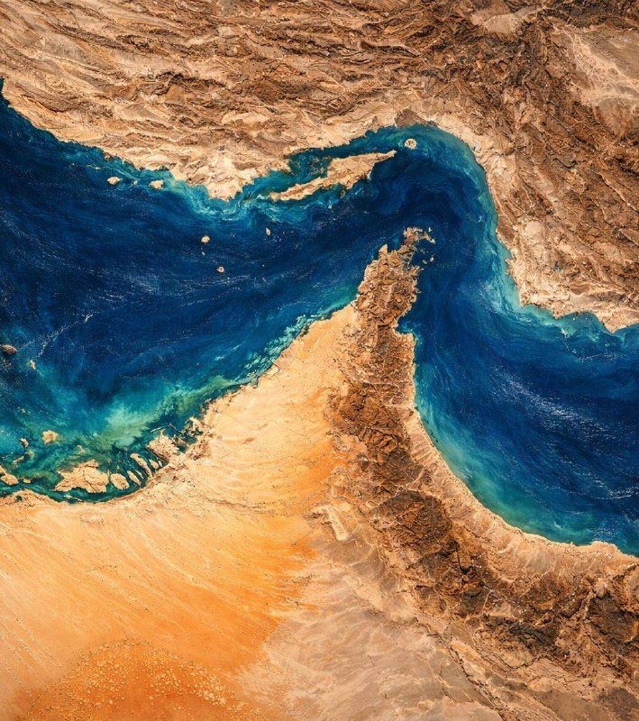 Satellite view showing a narrow, rugged landmass separating two bodies of turquoise and deep blue water, surrounded by arid tan and brown terrain.